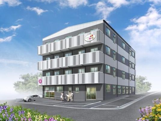 白とグレーの建物は３階建てで敷地内には駐車場があり、奥には緑があり、手前には花が咲いている。