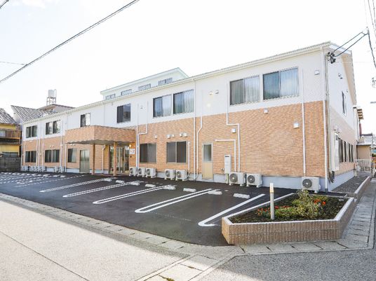 2階建ての建物は1階部分がベージュ、2階部分が白で、横に長く、敷地内には駐車スペースと小さな花壇もある。玄関には屋根が付き、日当たりも良好。