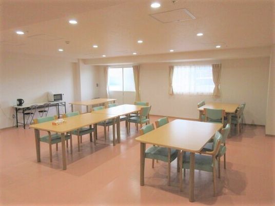 食堂には大きな窓が二つあり、木製の大きなダイニングテーブルが複数置かれ、壁際のテーブルには電子レンジが置かれている。