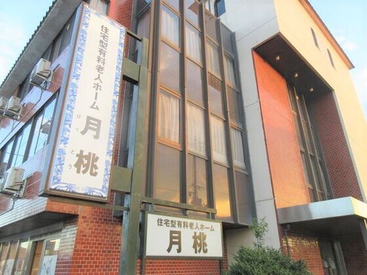 赤っぽいレンガ調の外壁に窓が多く付いた建物。敷地内には緑が植えられている。