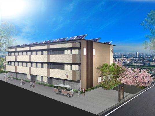 ベランダ付きの3階建ての建物の周りには桜の木があり、屋上にはソーラーパネルが設置されている。