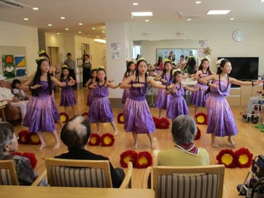 紫色のフラダンスを踊る女の子を椅子に座って眺める入居者たち。