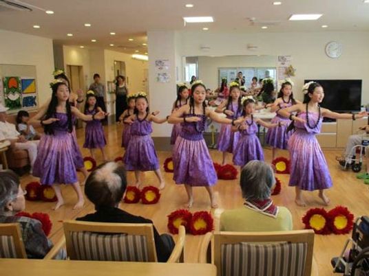 紫色のフラダンスを踊る女の子を椅子に座って眺める入居者たち。