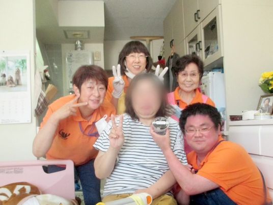 ピースサインをしている高齢女性を囲み微笑んでいるオレンジのポロシャツを着た男女のスタッフ。