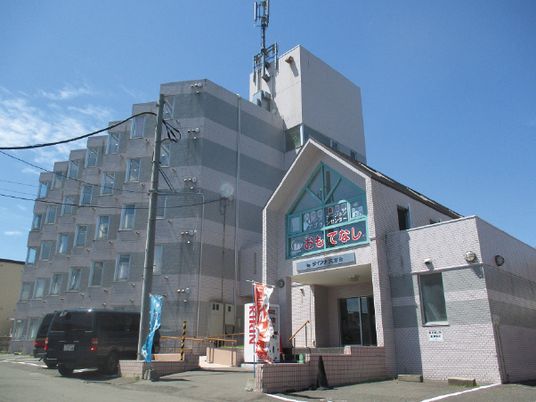 おもてなしと赤い文字で書かれた玄関先にはのぼりが立ち、奥には各フロアにベランダがあるグレーの建物がある。