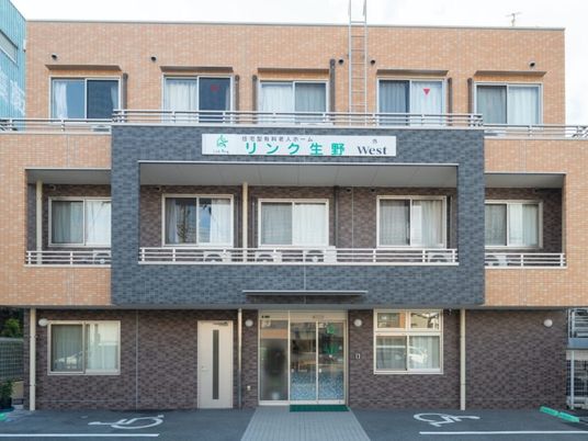 ブラウン系の外壁の3階建ての建物の前には駐車場があり、各フロアにベランダもある。