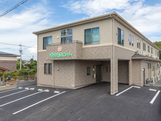 ベージュを基調とした2階建ての建物の外壁にはホーム名が書かれ、駐車場も付いている。
