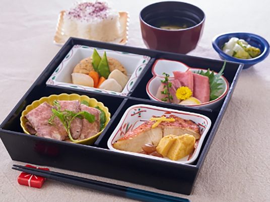 仕切りのついた弁当箱には魚料理や肉料理、刺身や煮物が盛られ、その周りには吸い物やゆかりご飯などもある。