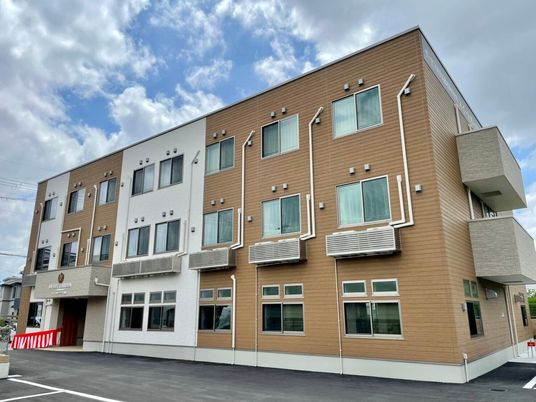 3階建ての建物はブラウンと白でまとめられていて、窓が多く、側面にはベランダもある。玄関には紅白の幕が設置され、駐車スペースもある。