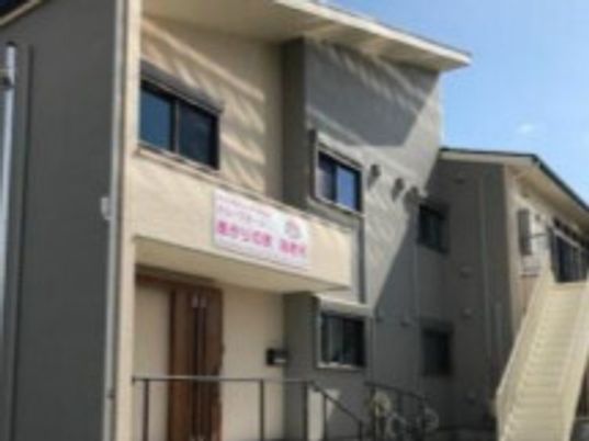 ベージュとグレー系の外観の２階建ての建物は日当たりがよく、玄関には屋根があり、手すり付きの階段が設置されている。