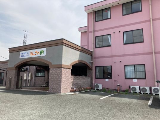 右側にはピンクの3階建ての建物があり、左側にはブラウン系の平屋建ての建物があり、駐車場もある。