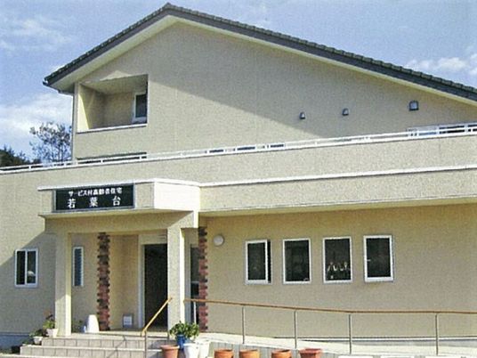 ベージュを基調とした2階建ての建物の外壁にはホーム名が書かれ、手すり付きスロープも付いている。