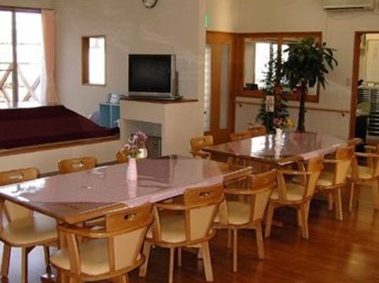四人掛けのテーブルが複数と椅子が並んでいる施設内のリビング。奥のほうにキッチンとリラックススペースがある様子