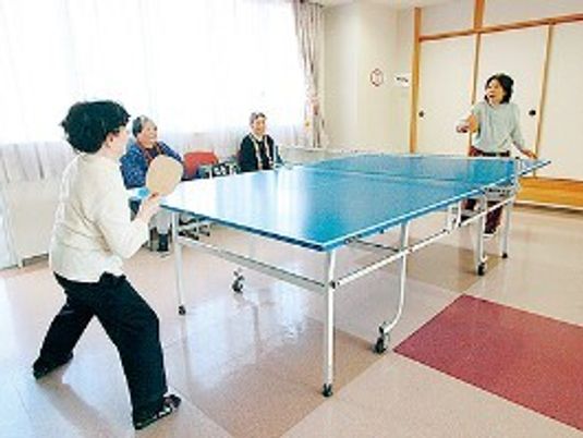 卓球台が置かれたスペースで女性２名が卓球をし、窓際で２名が見物をしている。大きな窓があり明るい雰囲気。