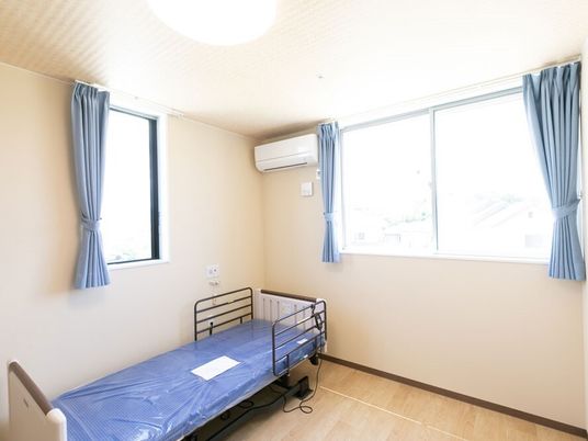 カーテンが付いた窓がたくさんあるフローリングの部屋。介護ベッドがあり、ベッドサイドには緊急通報装置もある。