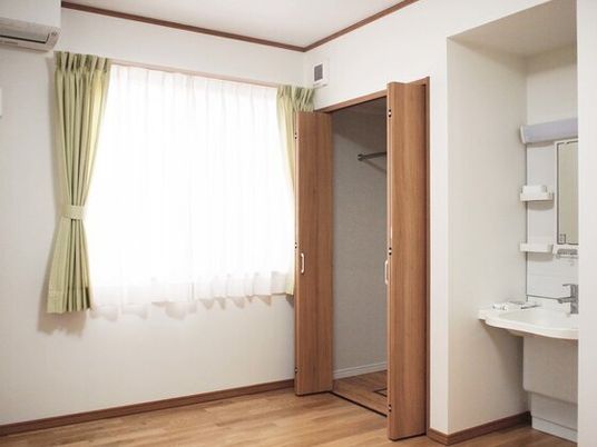 車いす対応の洗面所や大きなクローゼットが付いたフローリングの部屋。窓にはカーテンが付けられ、壁にはエアコンも設置されている。