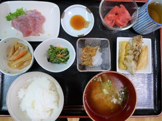 白いご飯に味噌汁、野菜の煮ものやあえ物、刺身にスイカ、お茶が入った湯飲みなどが並んだ黒いトレー。品数が多く食べ応えのある内容。
