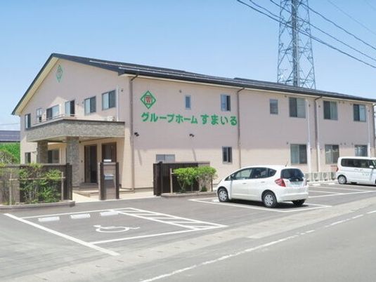 2階建ての建物は横に長く外壁には緑の文字でホーム名が書かれ、大きな駐車場も完備されている。玄関には屋根があり、建物周辺には緑も植えられている。