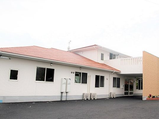 白い壁にオレンジ系の屋根が付いた２階建ての建物。玄関には車寄せが付き駐車場もあり、花が植えられたプランターが置かれている。