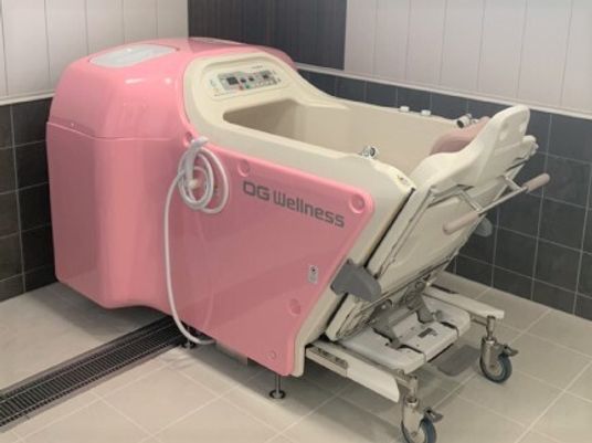 白とピンクの機械浴が置かれたタイル張りの浴室。機械浴の側面にはシャワーも付いている。