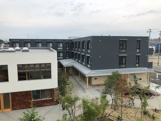 エル字型のグレーの建物は3階建て。そばには白い建物があり、周辺には木が植えられている。