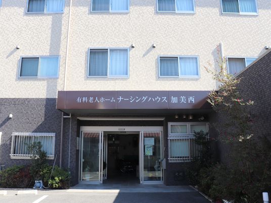 ３階建ての建物は白とベージュで玄関の屋根の部分に住宅名が書かれ、周辺に緑がある。