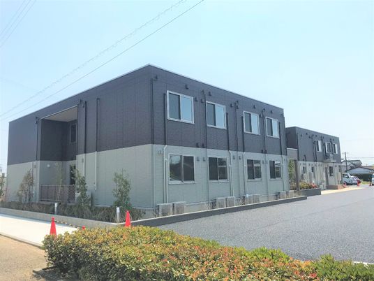 広い駐車場が付いた2階建ての建物。建物の周辺には緑が多くある。