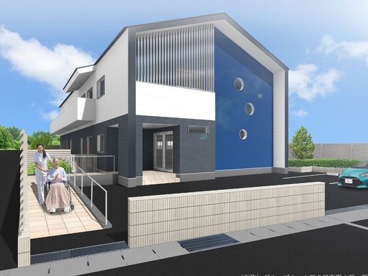 白とグレー、ブルーでまとめられた２階建ての建物。玄関から手すり付きのスロープがあり、車いすの女性にスタッフが付き添っている。