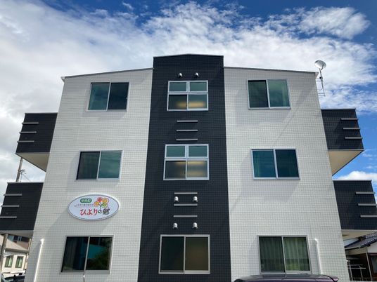 モノトーンの3階建ての建物は住宅地の中にあり、各フロアにベランダが付き、外壁には住宅名が書かれたプレートが貼られている。