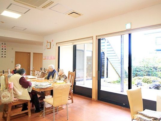 4名の女性入居者が木製テーブルを囲み、それぞれ好きなことをしている。大きな窓があり明るい雰囲気のリビングダイニング。