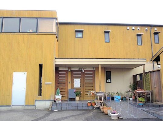 木目調の外壁が印象的な2階建ての建物。玄関には手すり付きのスロープがあり、たくさんのプランターやいすが置かれている。