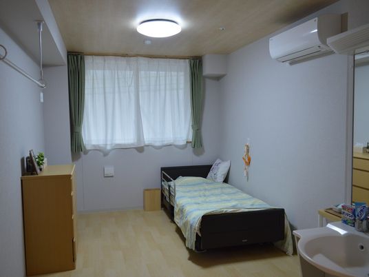 寝具付きのベッドやタンス、洗面所やエアコンなどがあるフローリングの部屋。大きな窓が付いている。