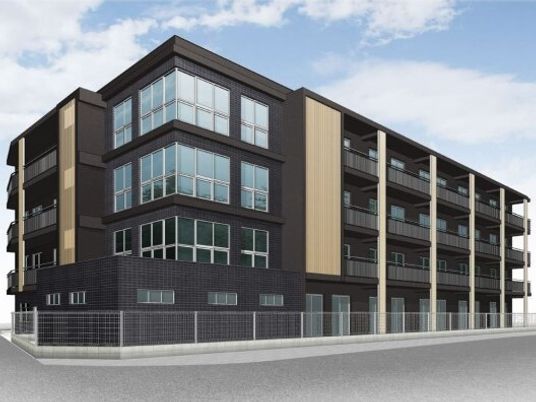 黒い外壁の4階建ての建物の角フロアにベランダがあり、建物の周りにはフェンスが設置されている。