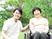 車いすの高齢女性と付き添いのエプロンを付けた男性スタッフ。緑の多い屋外で過ごしている。