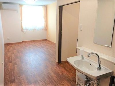 車いす対応の洗面所、が置かれたフローリングの部屋には大きな窓があり、カーテンが付いている。壁にはエアコンが付き、トイレもある。