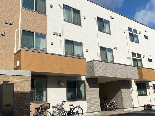 白とブラウンの３階建ての建物の前には自転車が止められている。日当たりのいい場所に建っている。
