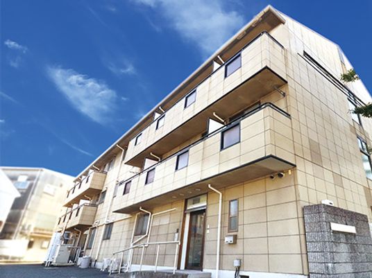 各フロアにベランダが設置された３階建ての建物。落ち着いたベージュ系でまとめられ、スロープも設置されている。