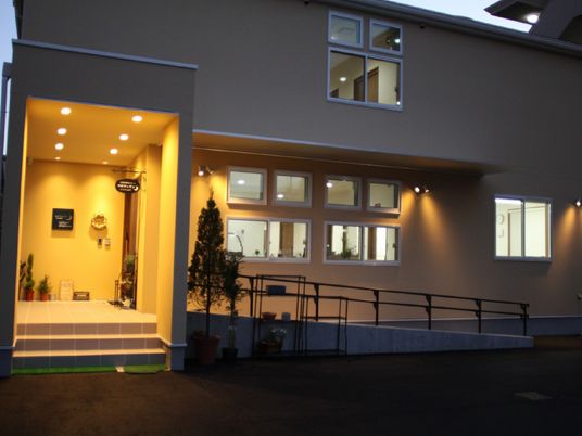辺りが薄暗くなり、玄関には明かりが灯り、たくさんの窓から明かりが漏れている２階建ての建物。