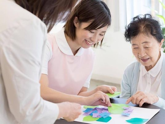 アジサイの貼り絵を楽しそうにしているピンクのエプロンを付けた若い女性スタッフと高齢女性。