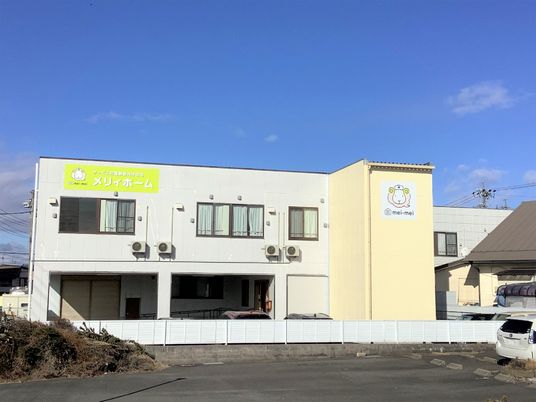 2階建ての建物は白を基調としていてベージュもアクセントとしてある。外壁にはヒツジのイラストが2か所描かれ、青い空が広がっている。
