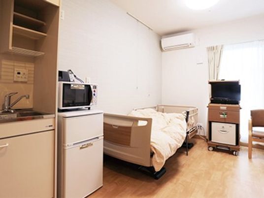 寝具付きのベッドが置かれ、エアコンやテレビ、キッチンが付いたフローリングの部屋には大きな窓が付いている。