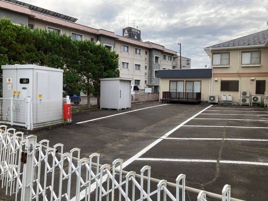 広い駐車場が付いた２階建ての建物はベージュの外壁でグレーの屋根があり、草木が植えられている。