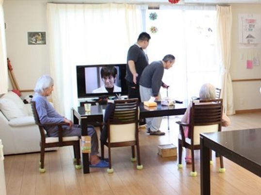 テレビのある広めのリビングでのんびりしている様子の高齢者とスタッフ。スタッフが一人の高齢者に寄り添っている