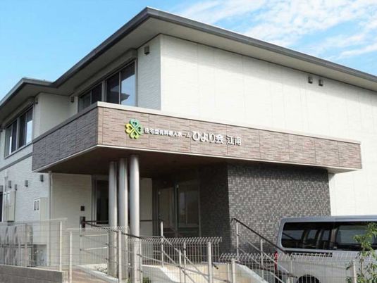 白い外壁の2階建ての建物にはホーム名が書かれたブラウンの屋根が付いていて、玄関周りには手すり付きの階段とスロープが付いている。