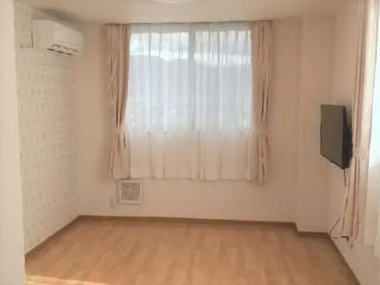 レースカーテンとカーテンが付いた大きな窓があるフローリングの部屋。エアコンと壁掛けテレビが設置され、白を基調としている。