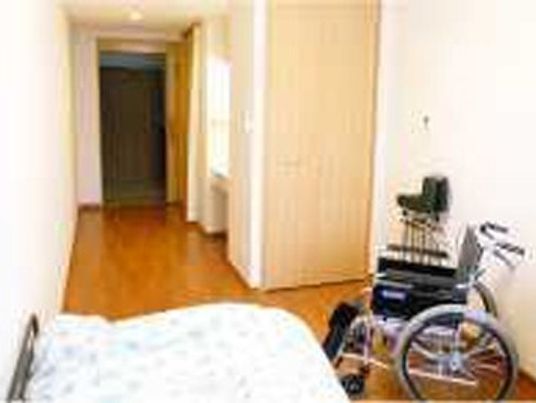 フローリングの部屋には寝具付きのベッドが置かれ、車いす対応の洗面所があり、車いすも置かれている。