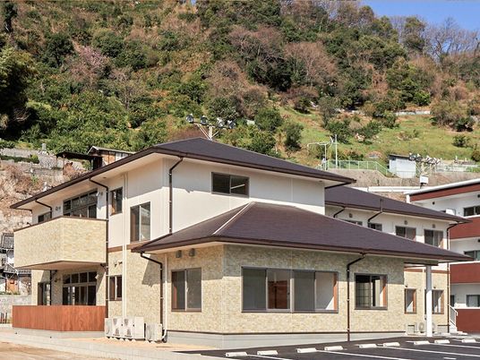 2階建ての建物にはグレーの屋根があり、後ろには山、周辺には住宅地があり、敷地内には駐車場がある。