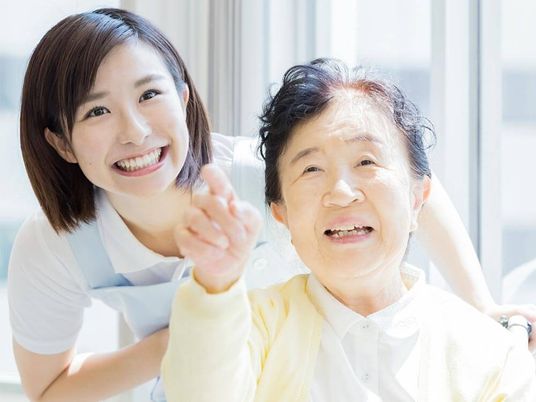 遠くを指さし笑っている車いすの高齢女性と一緒に見ているエプロンを付けた女性スタッフ。