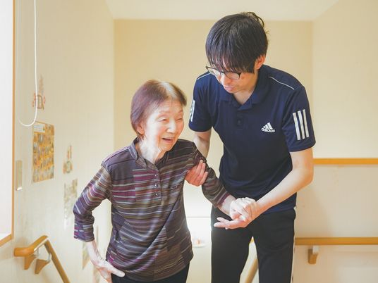 眼鏡をかけてポロシャツを着た男性スタッフが高齢女性の腕を支え、歩いている。