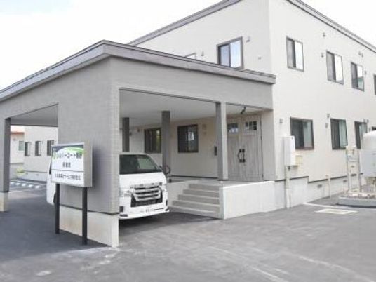 グレー系の色でまとめられた2階建ての建物の玄関には手すり付きのスロープと階段があり、車が止められている。